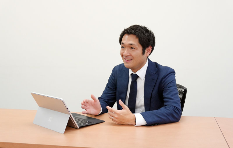 机に座り、タブレット端末を前に置いて身振り手振りを交えながら笑顔で話すスーツ姿の男性。