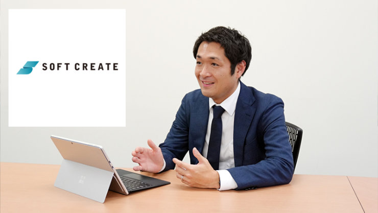 机に座り、タブレット端末の前で身振り手振りを交えて笑顔で話すスーツ姿の男性。左上にはSoft Createのロゴが配置されている。