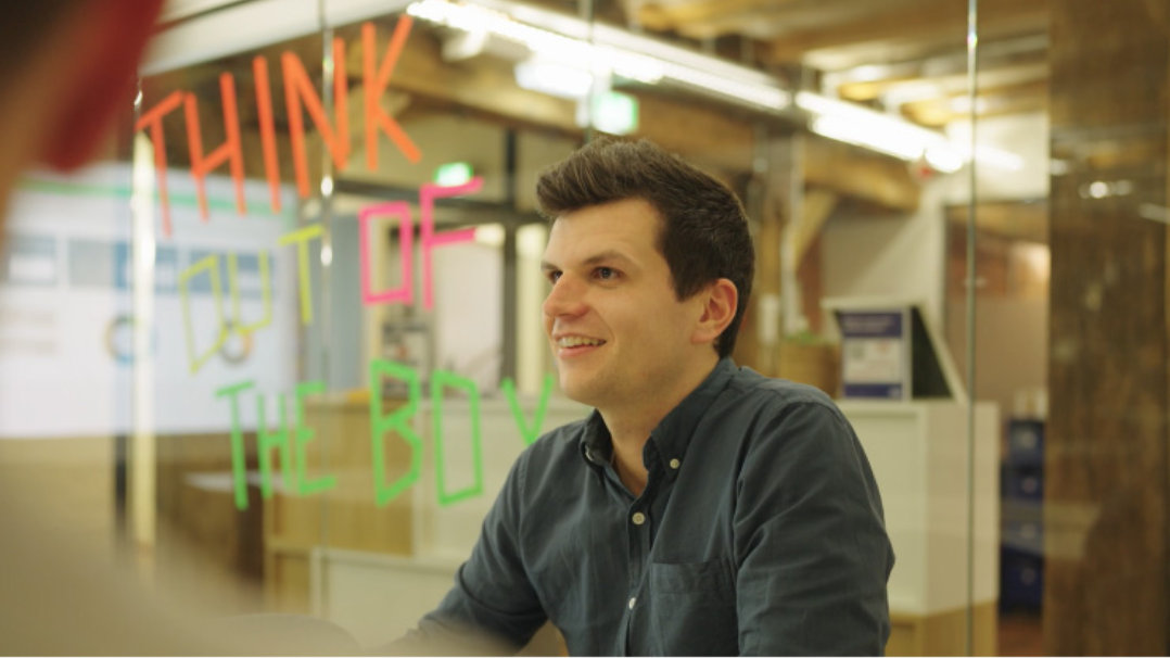 Eine Person sitzt in einem modernen Büroraum und blickt auf eine Glaswand, auf der in leuchtenden Farben die Worte „Denk um die Ecke“ geschrieben stehen.