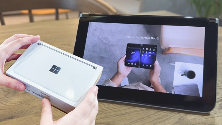Surface Duo 2 が画面に表示されたタブレットと手に持った Surface Duo 2 の箱