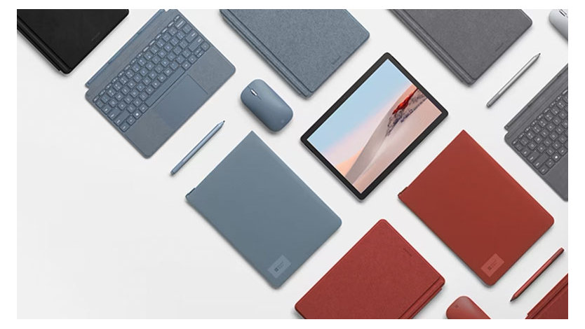 Surface Go 2のデバイスとアクセサリーが白い表面に配置されており、青や赤などさまざまな色のキーボード、ペン、マウス、保護ケースが含まれています。1台のデバイスには砂漠の風景が表示されています。
