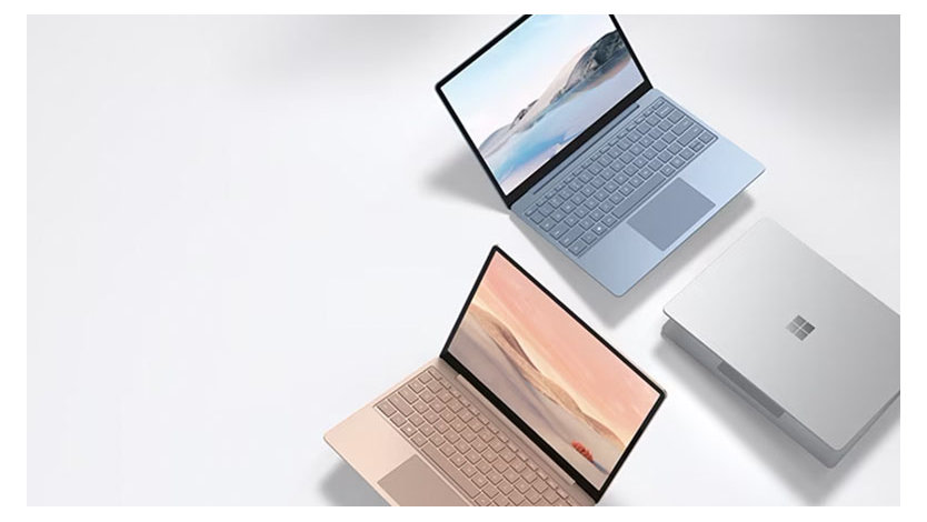 Here’s the translation to Japanese:  「2台のオープンなSurface Laptop Goデバイス、1台はライトブルー、もう1台はライトピンクで、Windows 11のスタート画面が表示されています。Microsoftロゴが入った閉じた白いSurface Laptop Goケースも横にあります。」