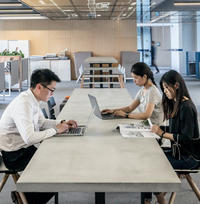 Three people seated at a long table working on laptops and reading a magazine