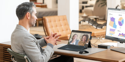 A person sitting at a desk participating in a Teams video call on a laptop connected to a desktop monitor displaying charts