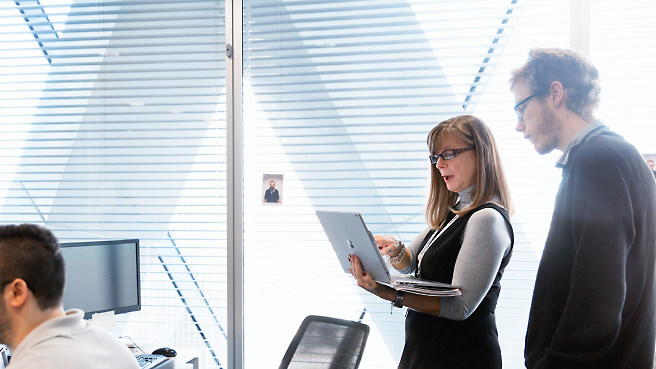 Dois profissionais analisando o conteúdo em uma tela de laptop em um ambiente de escritório moderno.