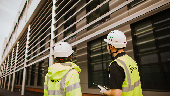Dois trabalhadores da construção civil, com coletes de segurança e capacetes, olham para um edifício moderno.