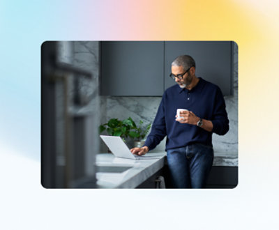 En person i en marineblå genser står på et moderne kjøkken, holder en kopp og ser på en bærbar datamaskin som står på benken.