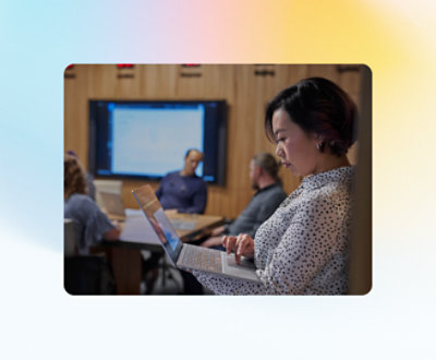 En kvinne står og bruker en bærbar datamaskin mens tre personer i bakgrunnen, som sitter ved et bord, fokuserer på en presentasjon.