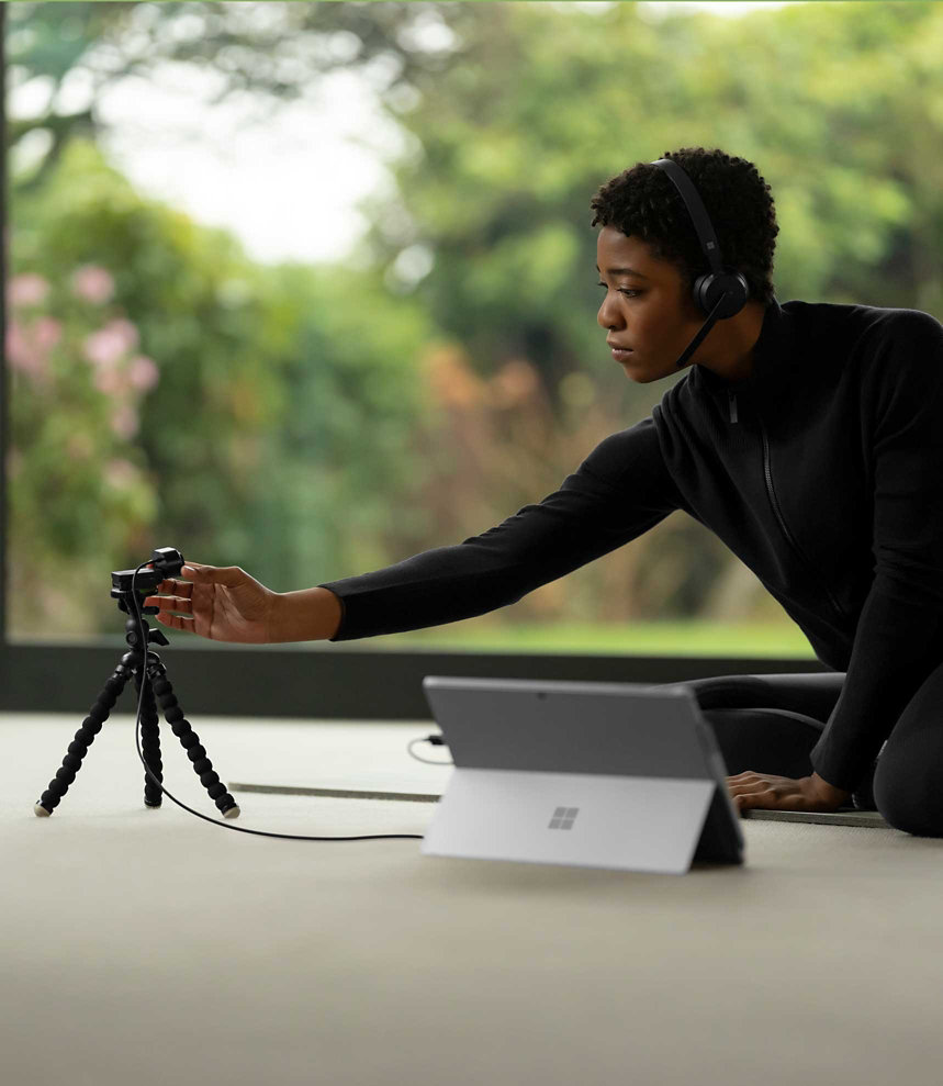 ผู้หญิงคนหนึ่งสวม Microsoft Modern Wireless Headset และโน้มตัวไปปรับ Microsoft Modern Webcam ที่อยู่ข้างแล็ปท็อป Surface Pro