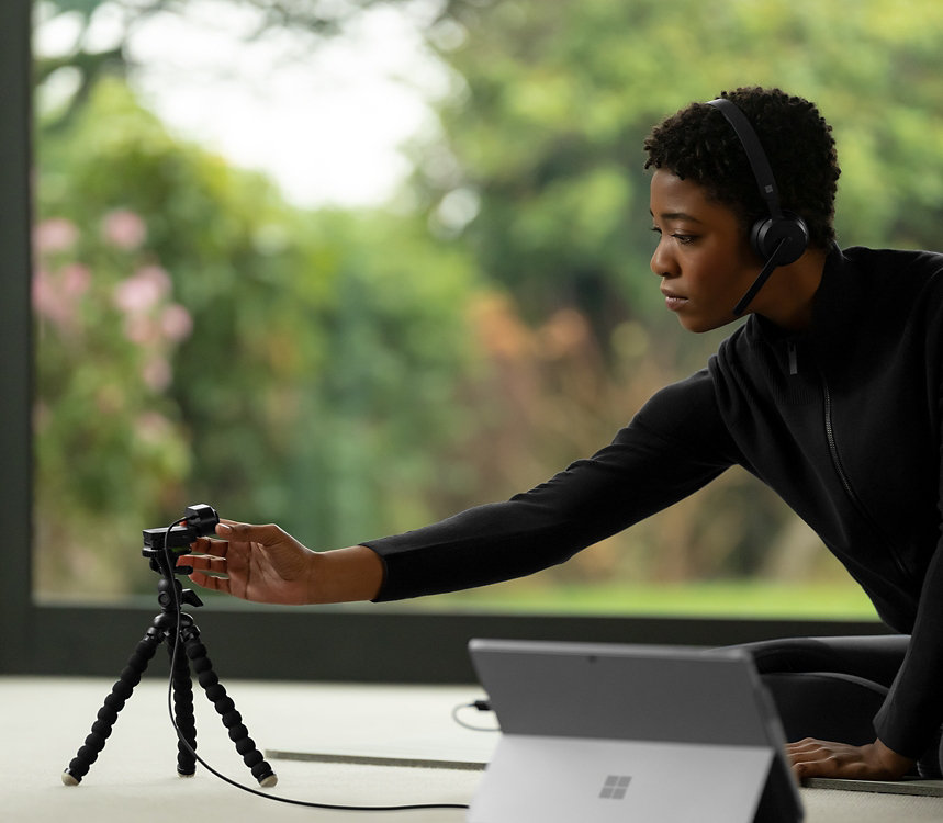 Une personne portant un casque sans fil Microsoft Modern et travaillant avec une caméra sur un trépied et un appareil Surface sur une table.