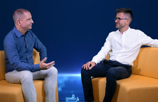 Das Bild zeigt Stratos und Sven, die sich gegenüber auf einem gelben Sofa sitzen vor einem blauen Hintergrund.