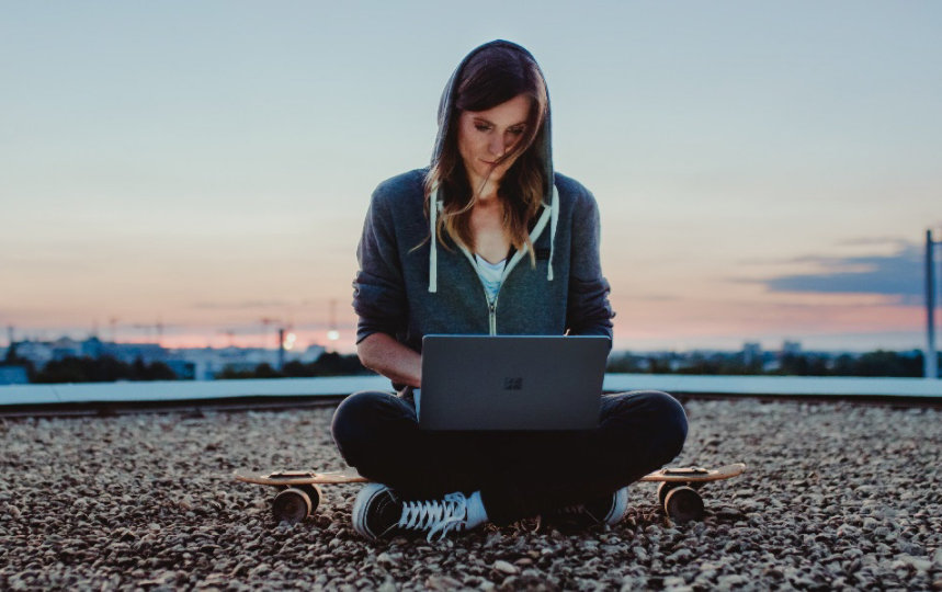 Eine Frau sitzt mit ihrem Skateboard und Laptop auf einem mit Kies bedeckten Dach.