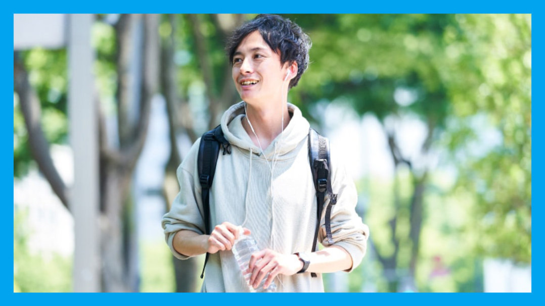 バックパックを背負った若い男性が水筒を持ち、笑顔で公園を歩いている。