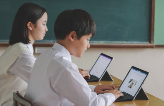 制服を着た 2 人の学生が机に座ってタブレットで作業しています