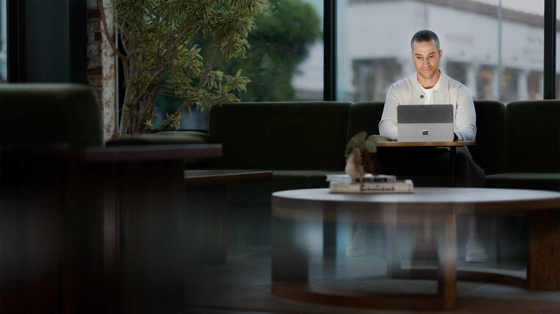 Office worker sitting in front of a large window with an open laptop, behind a coffee table with two books and a plant on it.