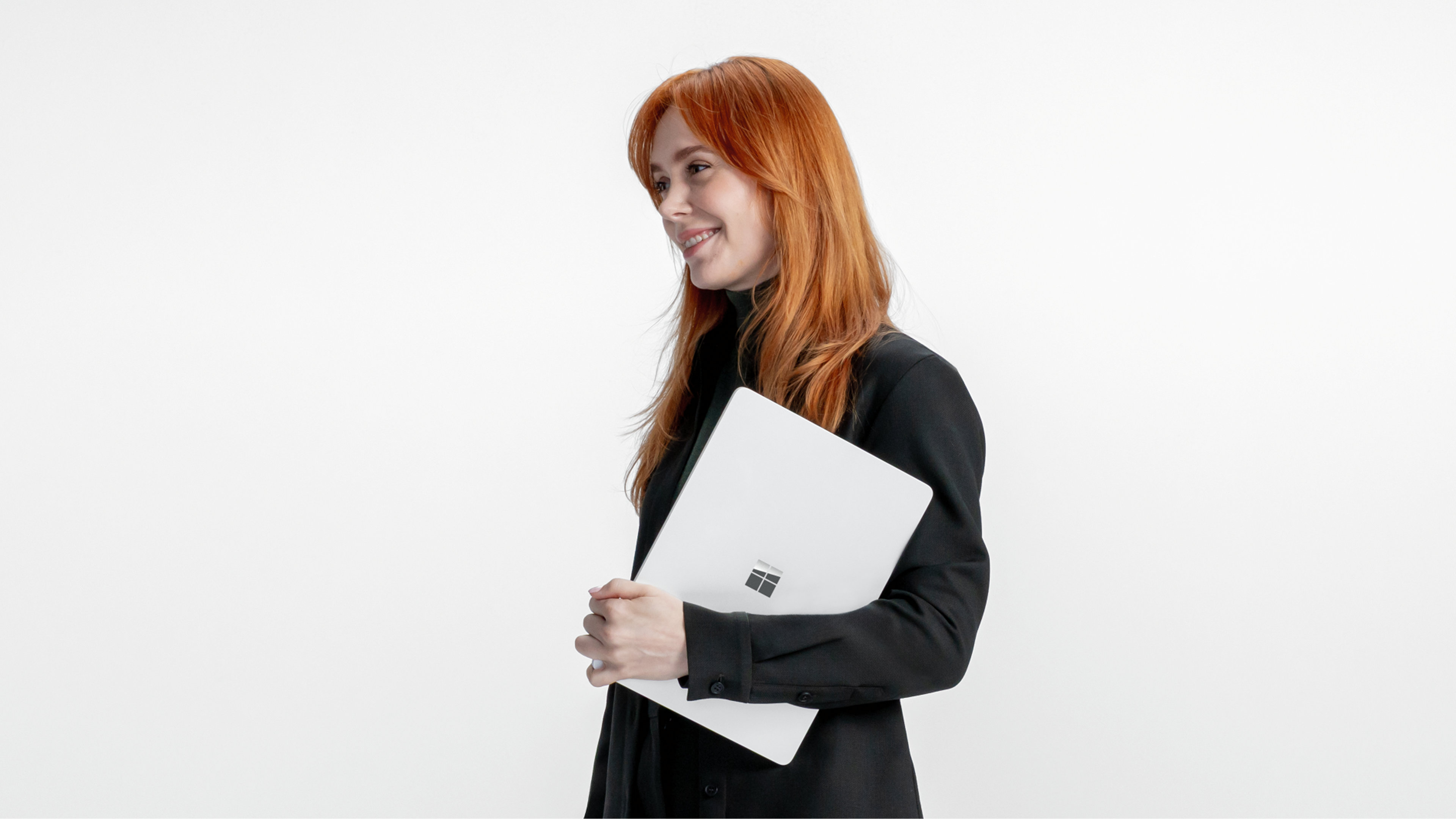Une personne souriante regarde vers la gauche en tenant un ordinateur portable Windows chrome fermé sur un arrière-plan gris
