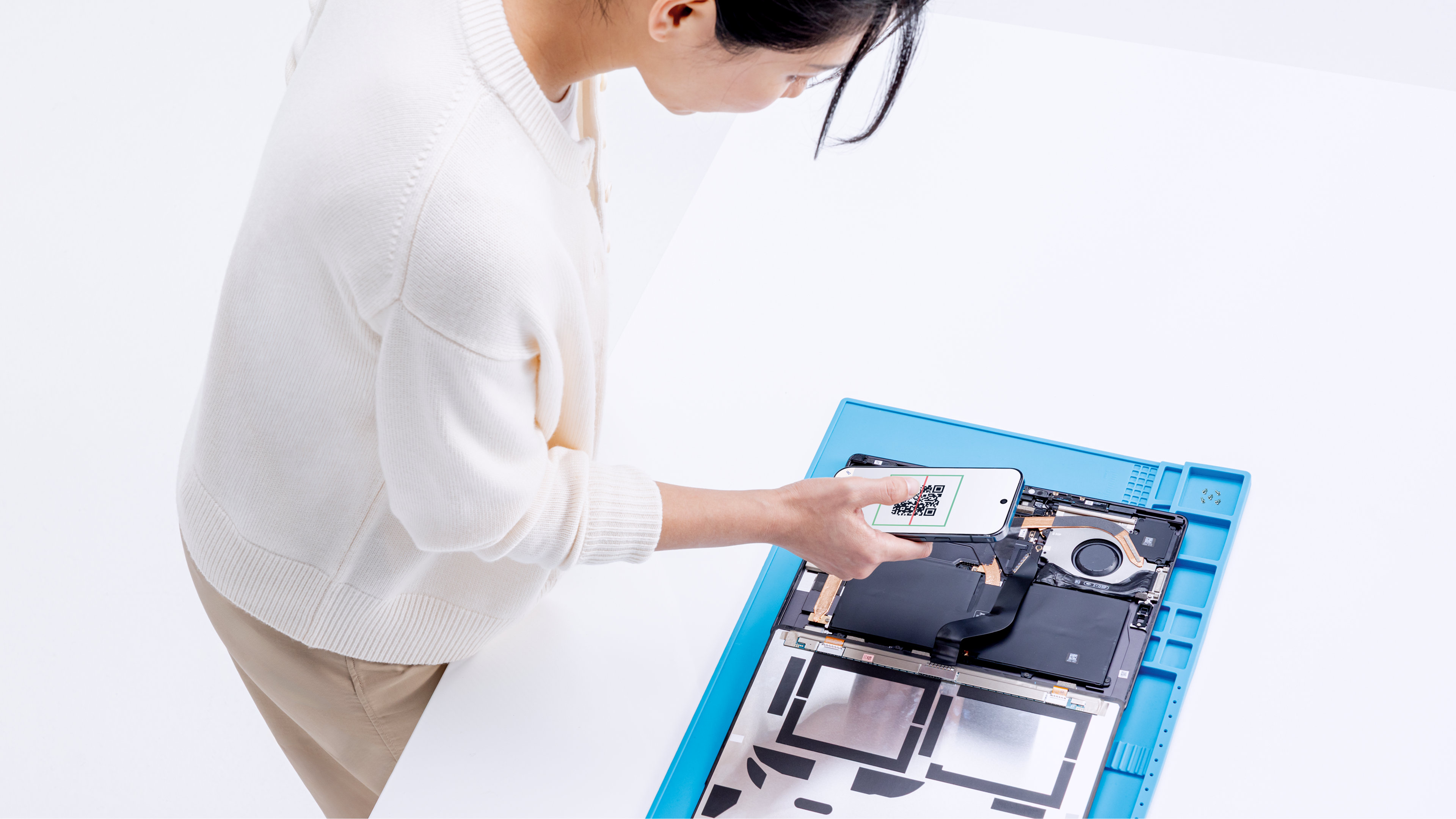 Un technicien informatique se tient au-dessus d'une vue de l'intérieur d'un ordinateur portable Windows sur un plateau bleu, à la recherche de pièces prêtes à être réparées, prenant une photo à l'aide d'un appareil mobile.
