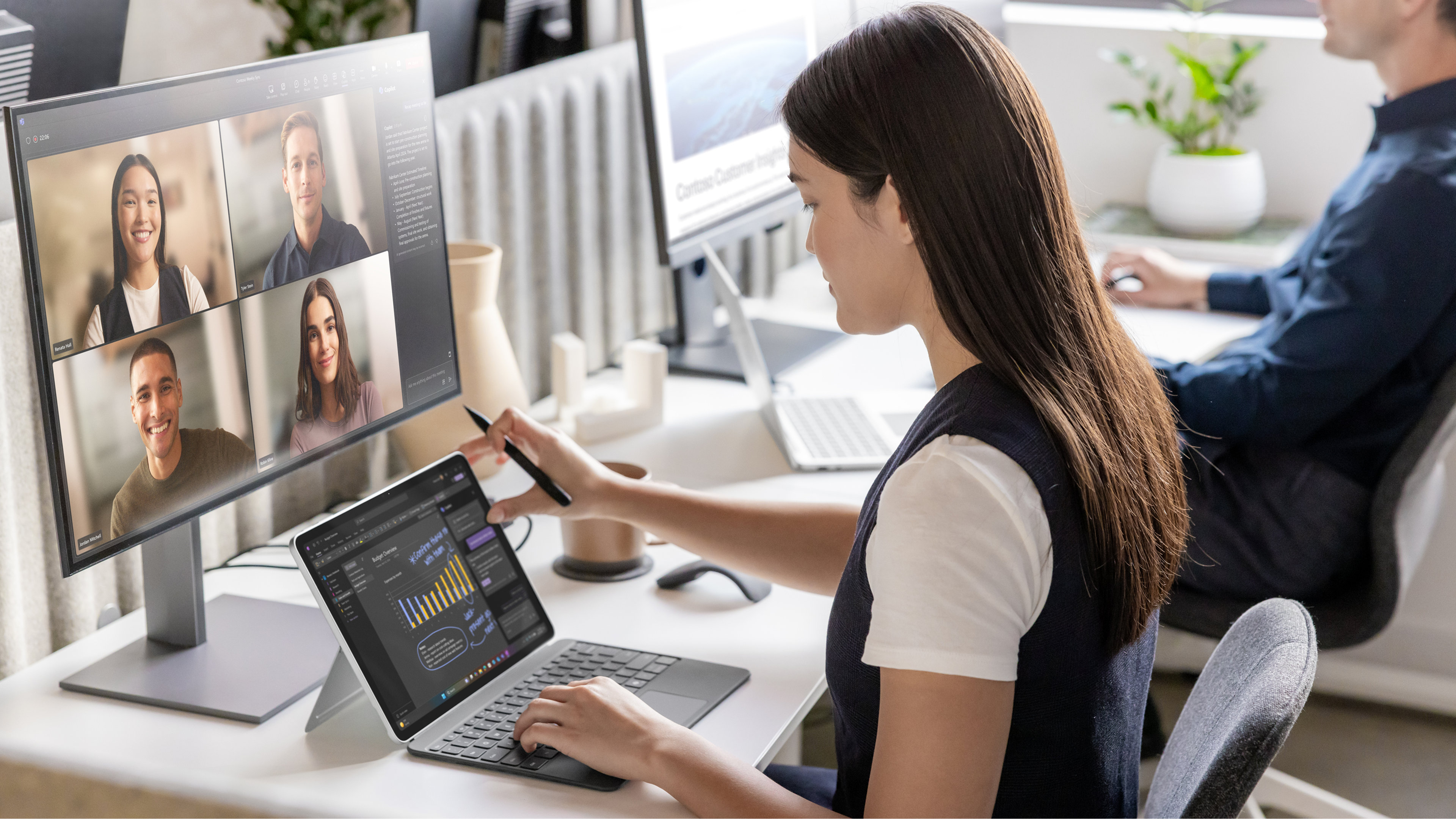 Deux employés de bureau assis à des postes de travail, l'un avec un ordinateur portable Windows ouvert et un grand écran externe, participent à un appel vidéo avec quatre autres employés, avec des tasses à café, des souris et des plantes à chaque bureau.