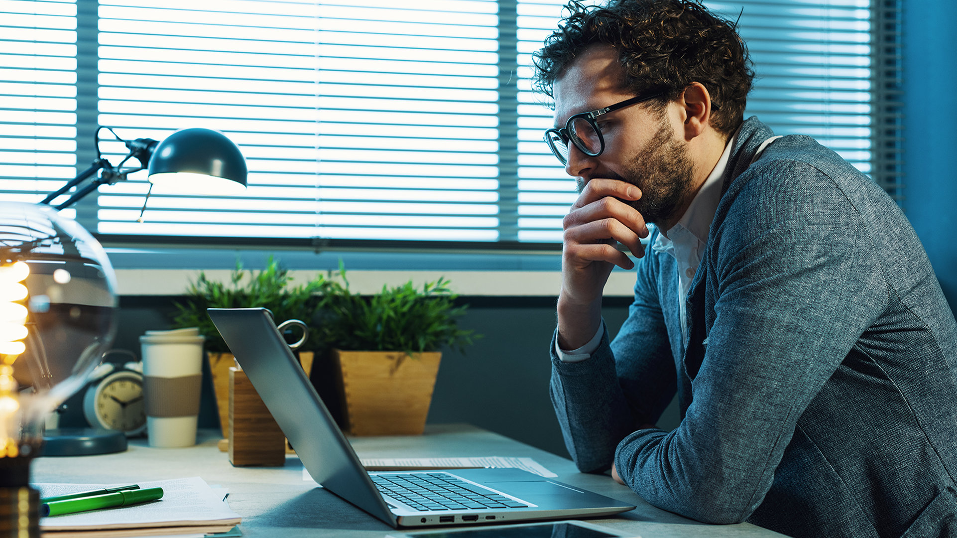 Office worker sitting at a desk beside a window with an open laptop, a mobile device, desk lamp, clock, coffee cup, and plants.