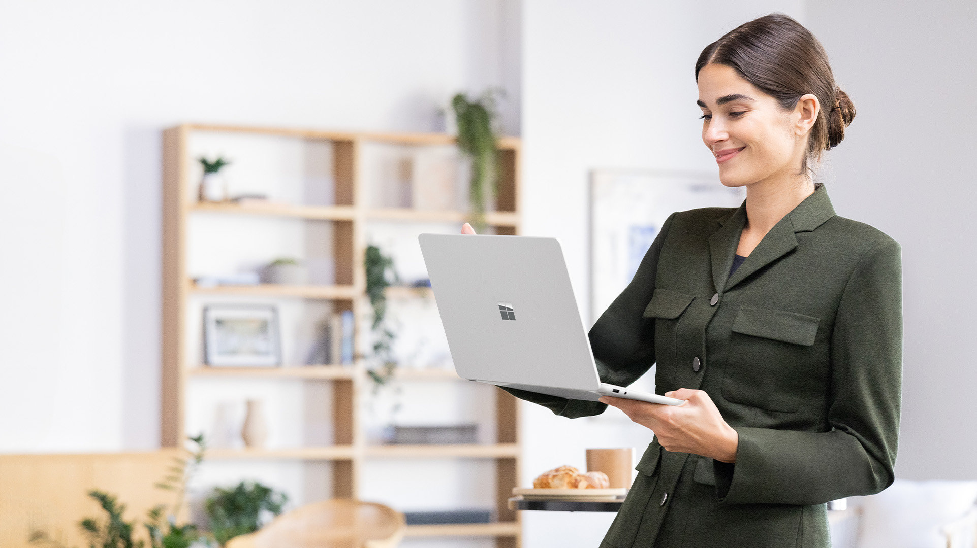 A remote worker standing and holding an open Windows 11 Surface for Business device with a table, chair, plants, and a bookshelf in the background.