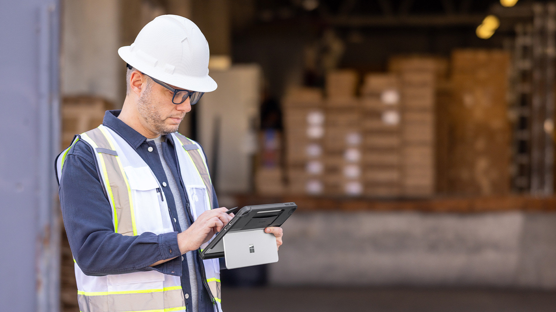 Warehouse worker wearing a safety vest and hard hat working on a Surface Pro with a loading dock in the background
