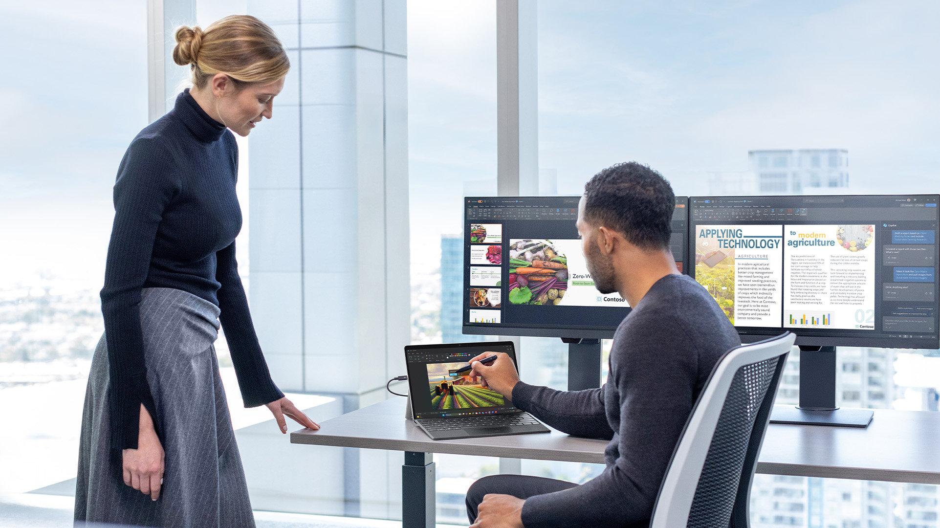 Two office workers, one sitting and one standing, at a workstation with an open Windows 11 Surface for Business device with a stylus, two external monitors, all displaying different webpages.