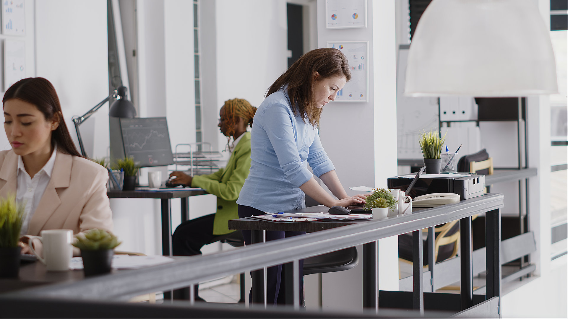 Three office workers, one standing and two seated, each at a workstation with laptops and miscellaneous office furniture, lamps, notebooks, and plants.