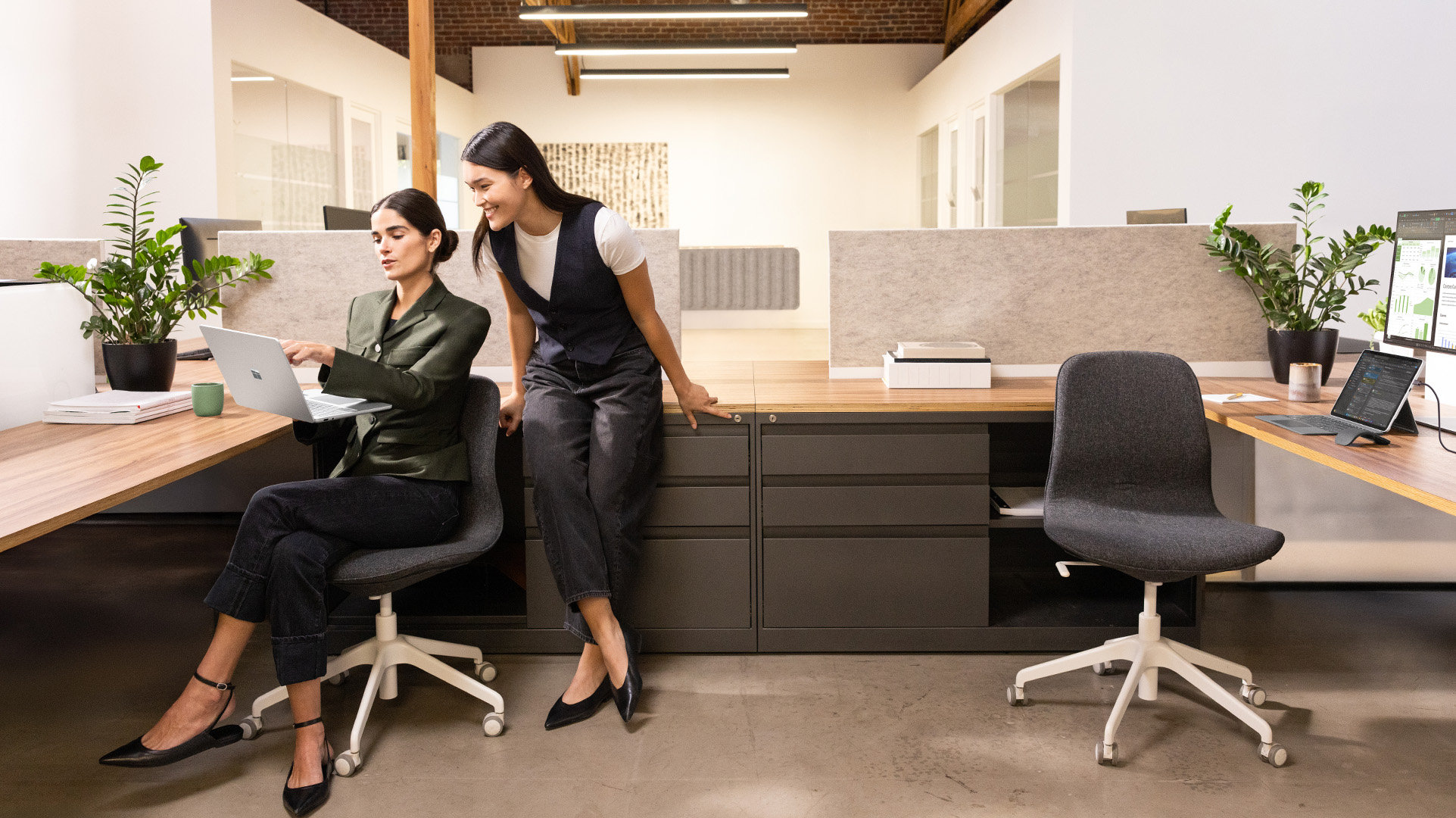 Two office workers in a large shared workspace, one seated and one standing, with Surface for Business devices discussing on-screen data.