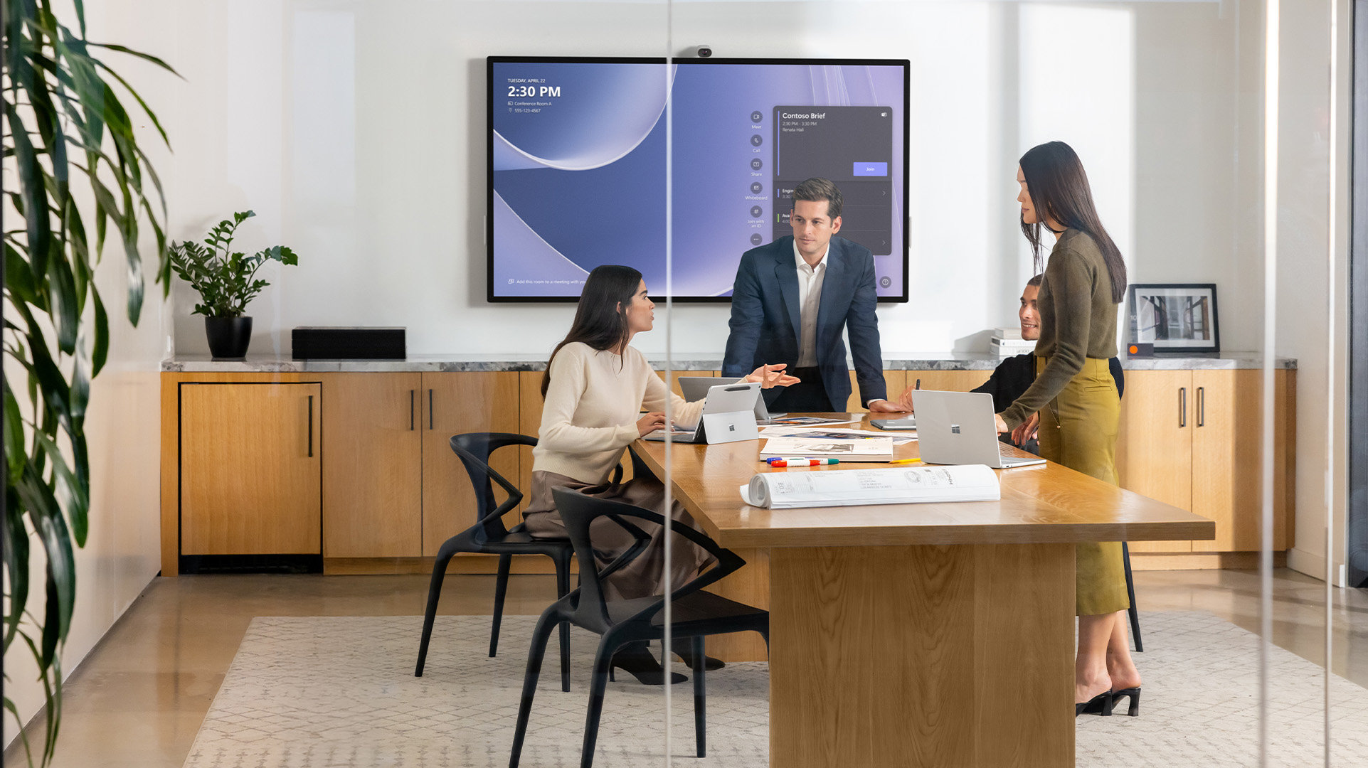 Four people meeting in a large conference room all using Surface for Business devices and a Surface Hub 3 mounted on the wall in the background over a row of built-in cabinets