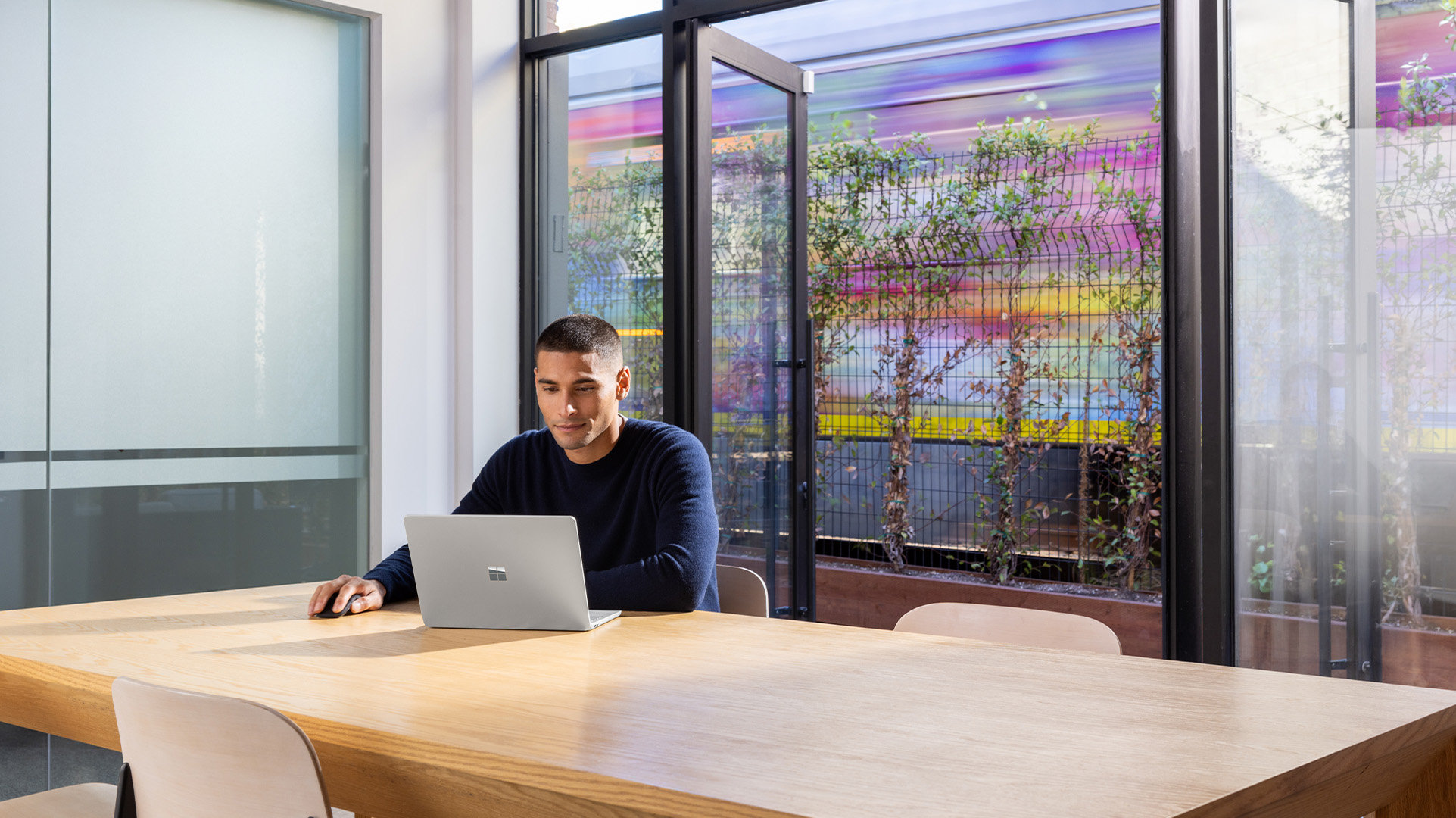 Office worker sitting alone in a meeting room with an open Windows 11 Surface for Business device, and doors in the background opening out into an open public space.