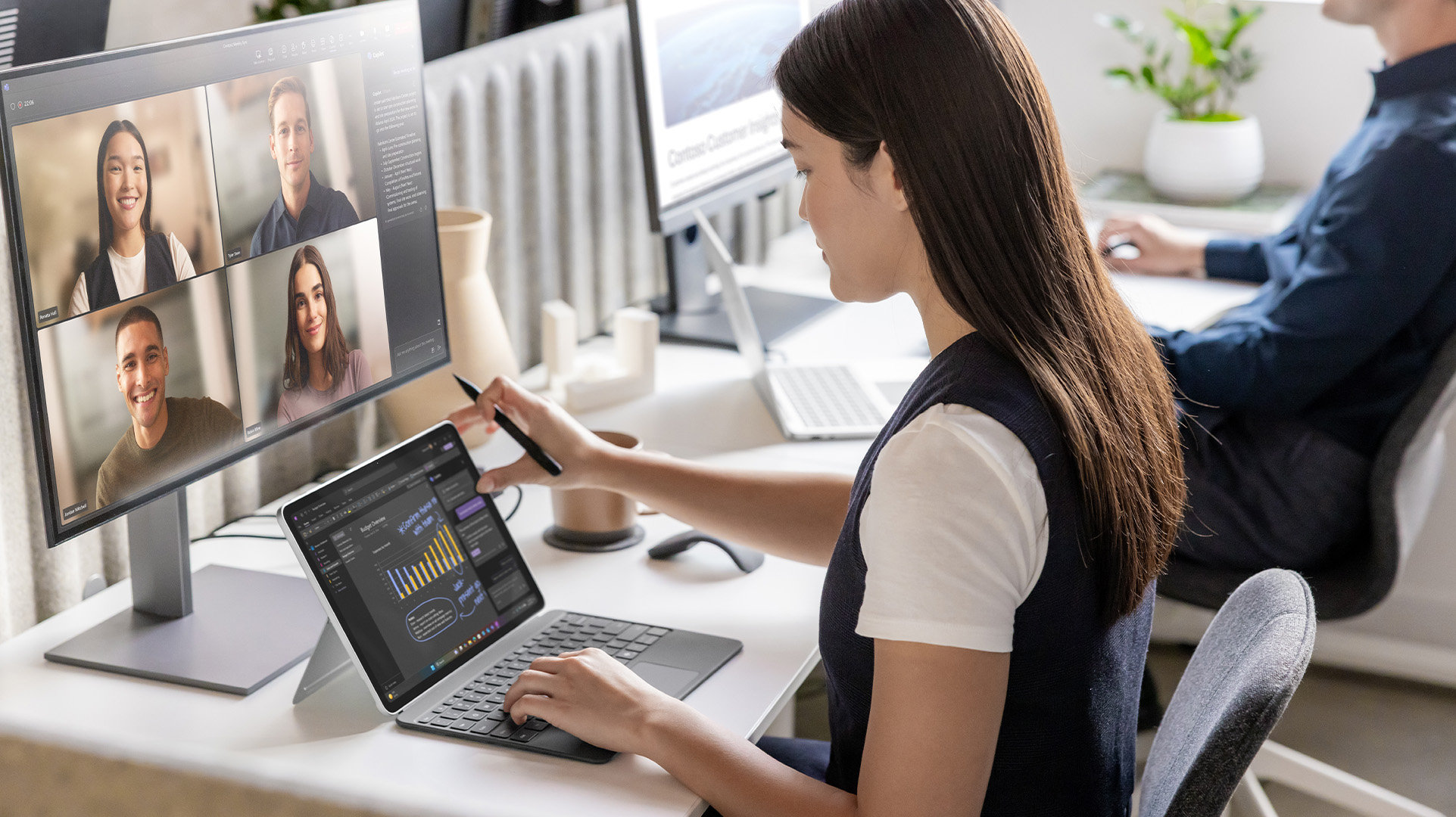 Two office workers sitting at a shared workstation, one using a Surface for Business device with an external monitor in an online meeting with four coworkers, the other in the background also working on a large external monitor.