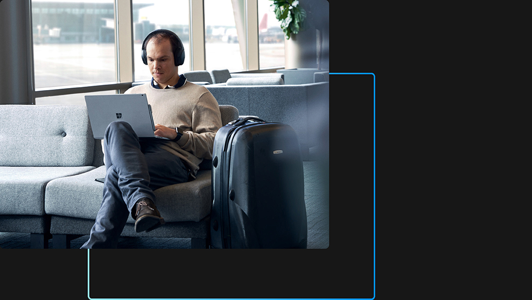Een persoon die in een luchthavenlounge zit met een koptelefoon op en zijn of haar laptop gebruikt met zijn of haar benen gekruist en zijn of haar koffer in de buurt
