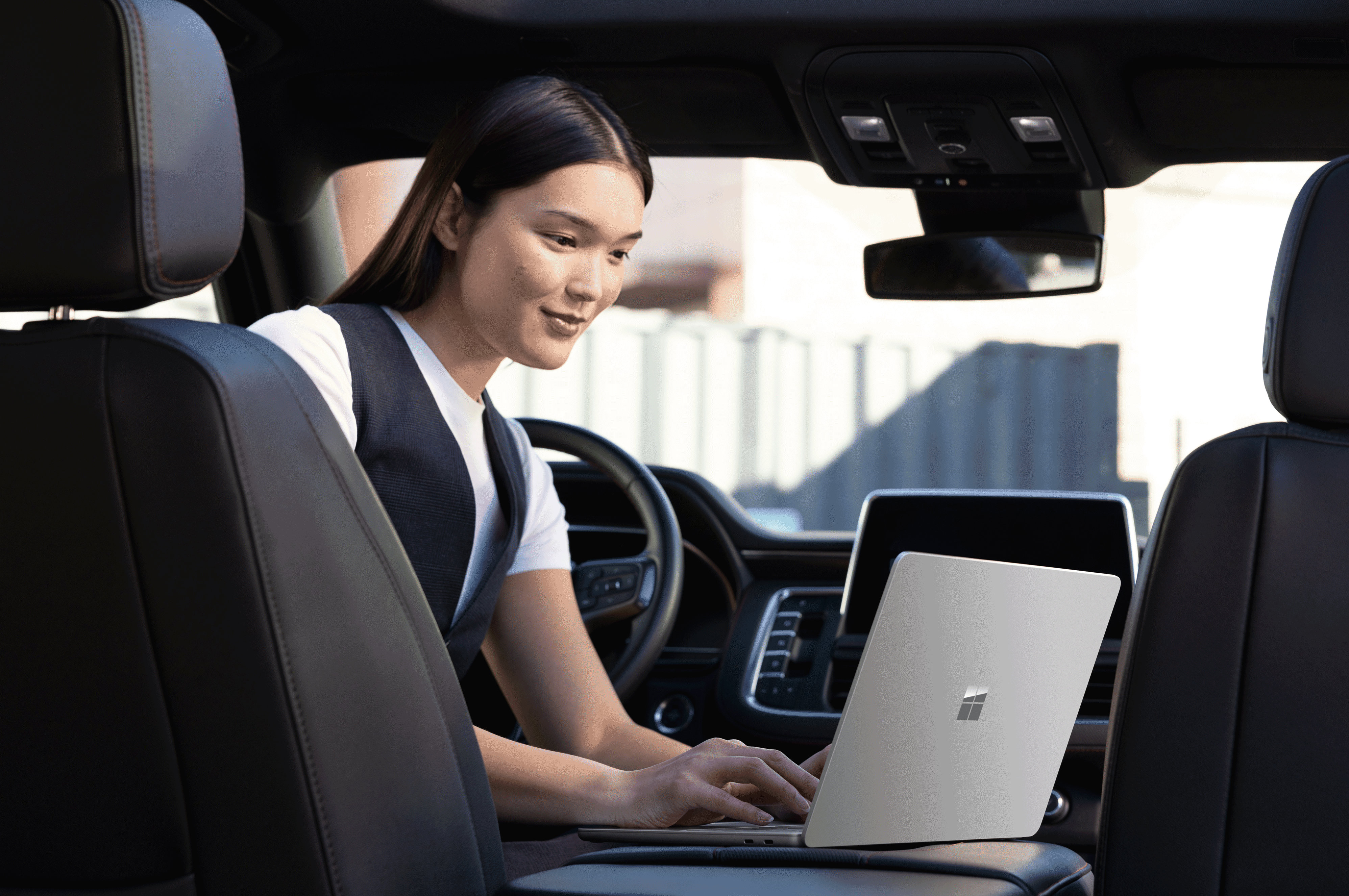 A person seated in the backseat of a car, working on a Surface laptop placed on their lap