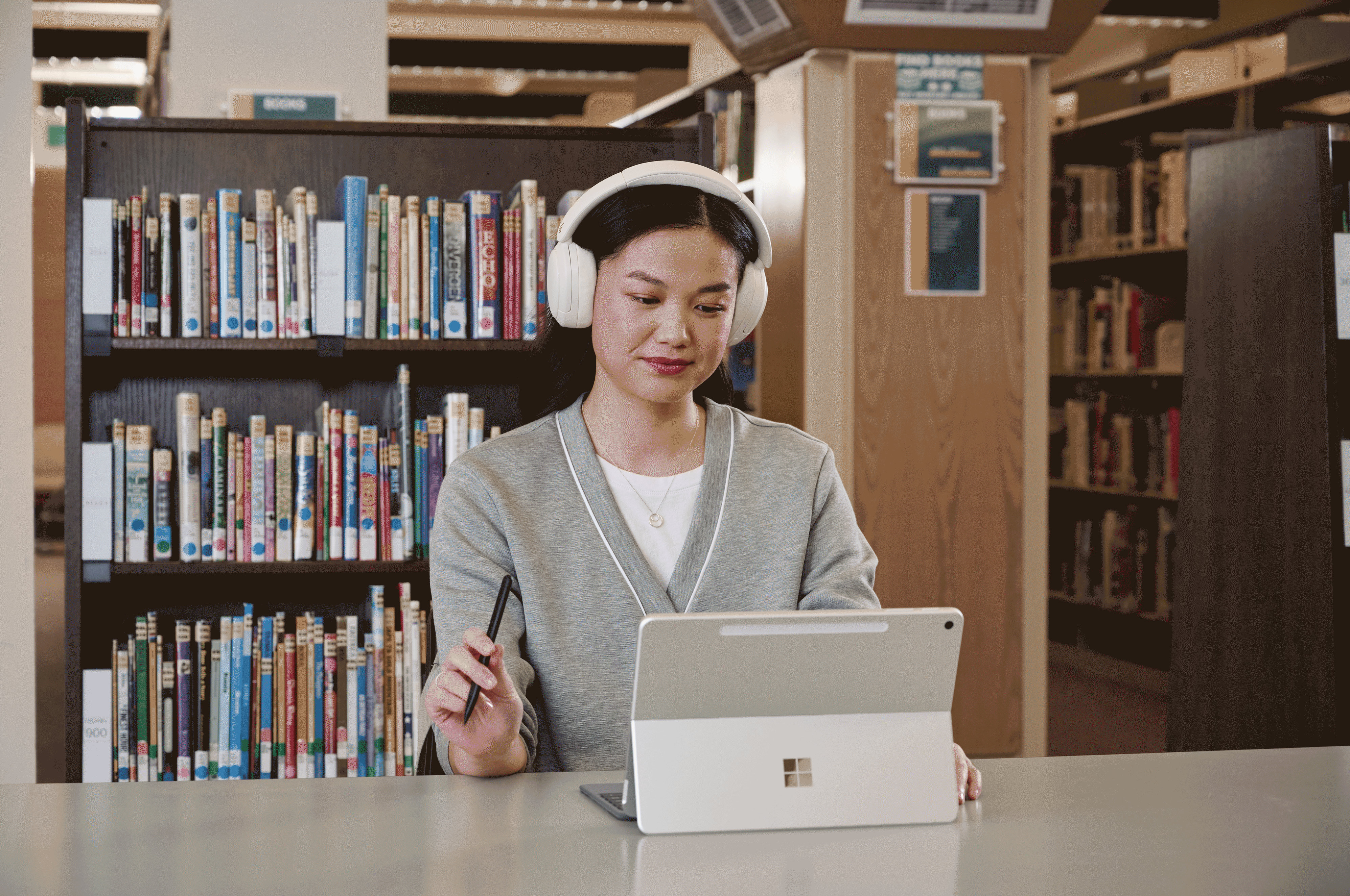 A person wearing large headphones and using a Surface device with a pen while seated at a table in a library