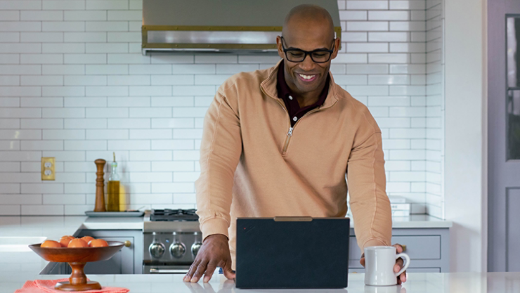 A person in a kitchen working on their laptop while smiling and gripping a mug