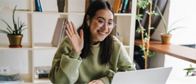 緑のセーターを着た、眼鏡をかけた人がノート PCの前のデスクに座り、笑顔で手を振っている。