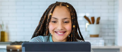 Ein lächelndes Kind mit zu Zöpfen geflochtenem Haar sitzt vor einem Laptop in einer küchenähnlichen Umgebung.