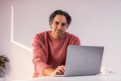 A man wearing glasses, sitting at a desk with a laptop, smiling.