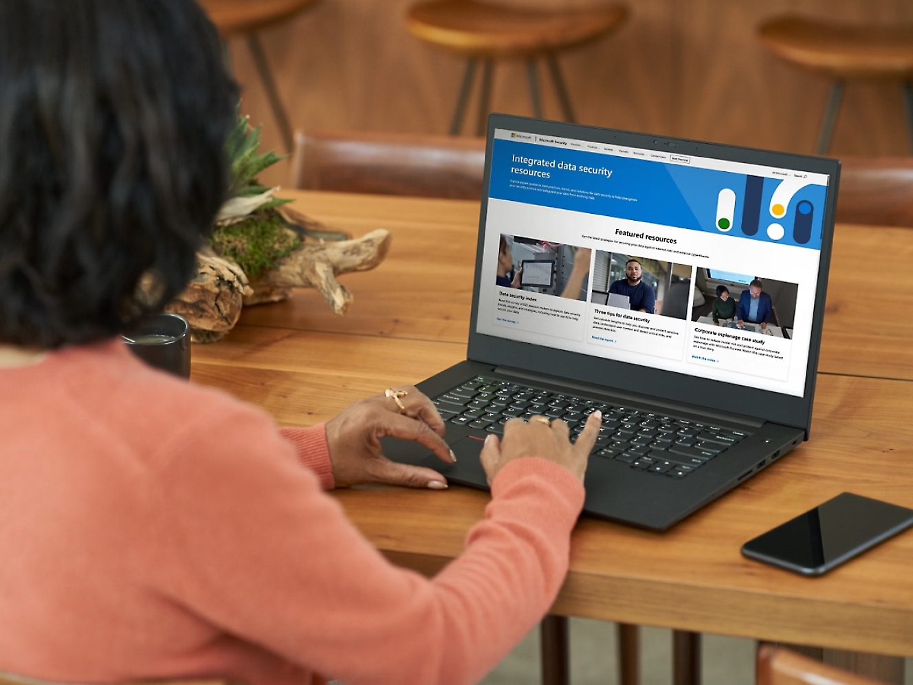 Mujer con jersey naranja usando un portátil que muestra en su pantalla un sitio web sobre seguridad de datos en una mesa de madera