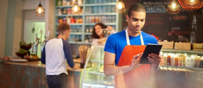 Barista con maglia blu e grembiule arancione lavora al tablet in un bar luminoso. Scaffali con vari prodotti 