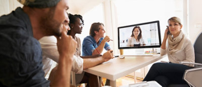 Quattro persone sedute a un tavolo guardano con attenzione lo schermo di un computer su cui è visualizzata una videochiamata con una persona.