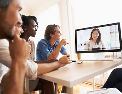 A group of people sits at a table, engaging in a video conference with a person displayed on a computer screen.