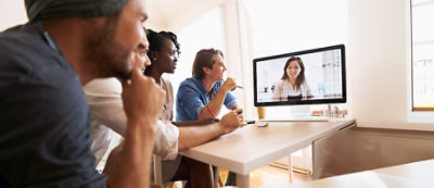 A group of people sits at a table, engaging in a video conference with a person displayed on a computer screen.