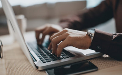 Osoba kuca na laptop računaru pored kojeg se na stolu nalazi pametni telefon.