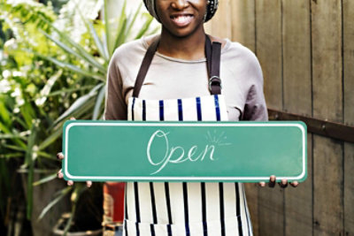 Osoba sa keceljom drži zeleni natpis „Otvoreno“. U pozadini se nalaze biljke.