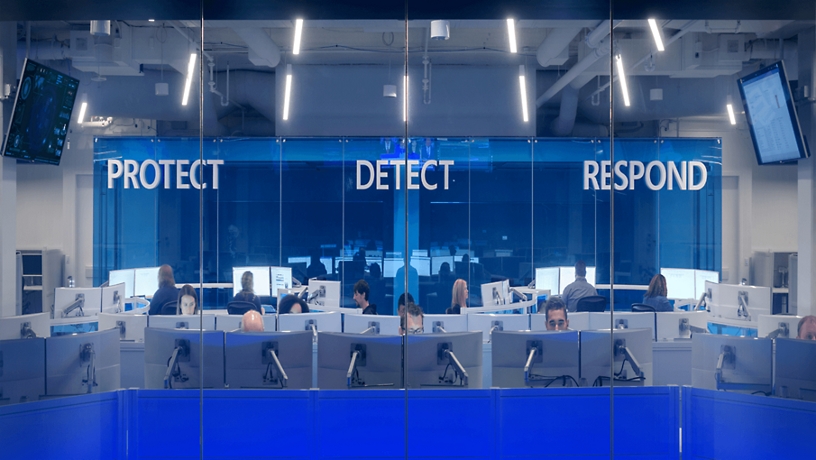 Personer, der arbejder ved skriveborde på et kontor, hvor ordene Beskyt Registrer og Reager står på væggen.