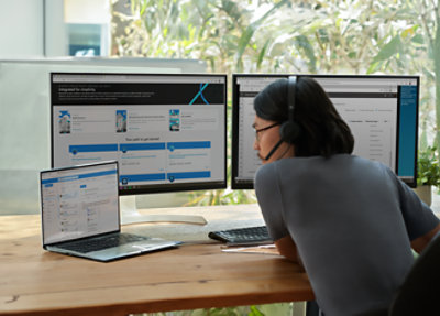 Person wearing headphones working at a desk with a laptop and two monitors displaying various applications.