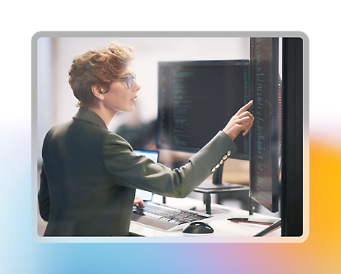 Une femme qui travaille avec plusieurs écrans affichant du code et qui pointe son doigt sur une partie du code
