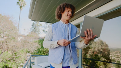 A person stands on a balcony holding an open laptop, with trees and a palm tree in the background.