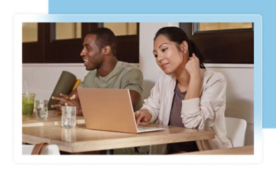 2 人がノート PC と飲み物を置いたテーブルに座っています。右側の人がノート PC を見ています。